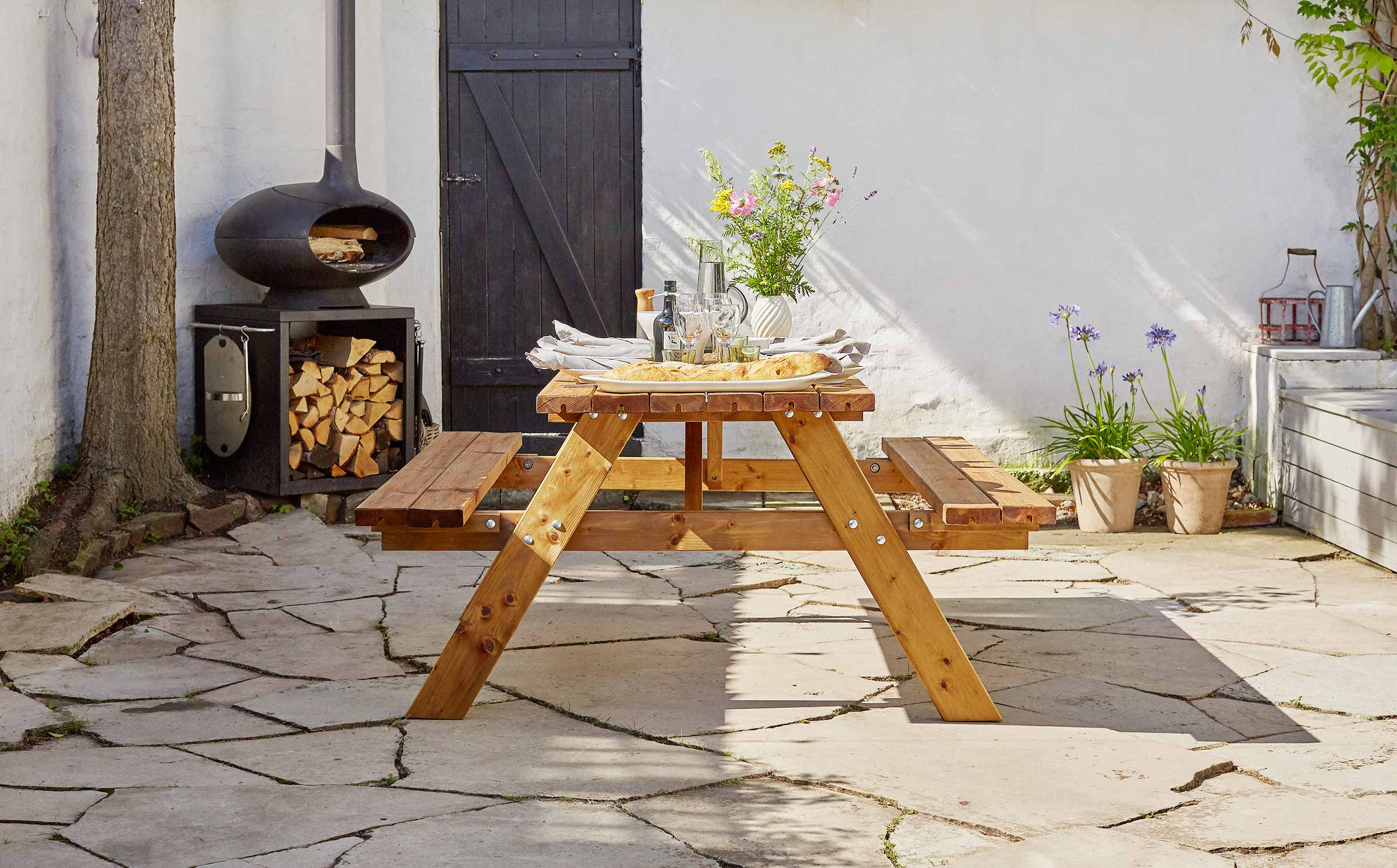 A brown A-frame picnic table with flowers and food, next to an outdoors fireplace