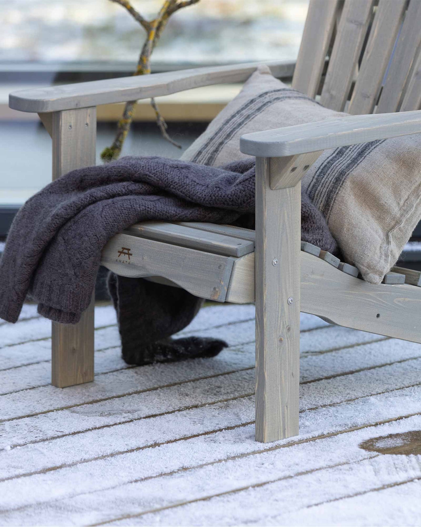 Closeup of a grey wooden Adirondack chair with a decorative throw pillow and blanket on a snowy patio