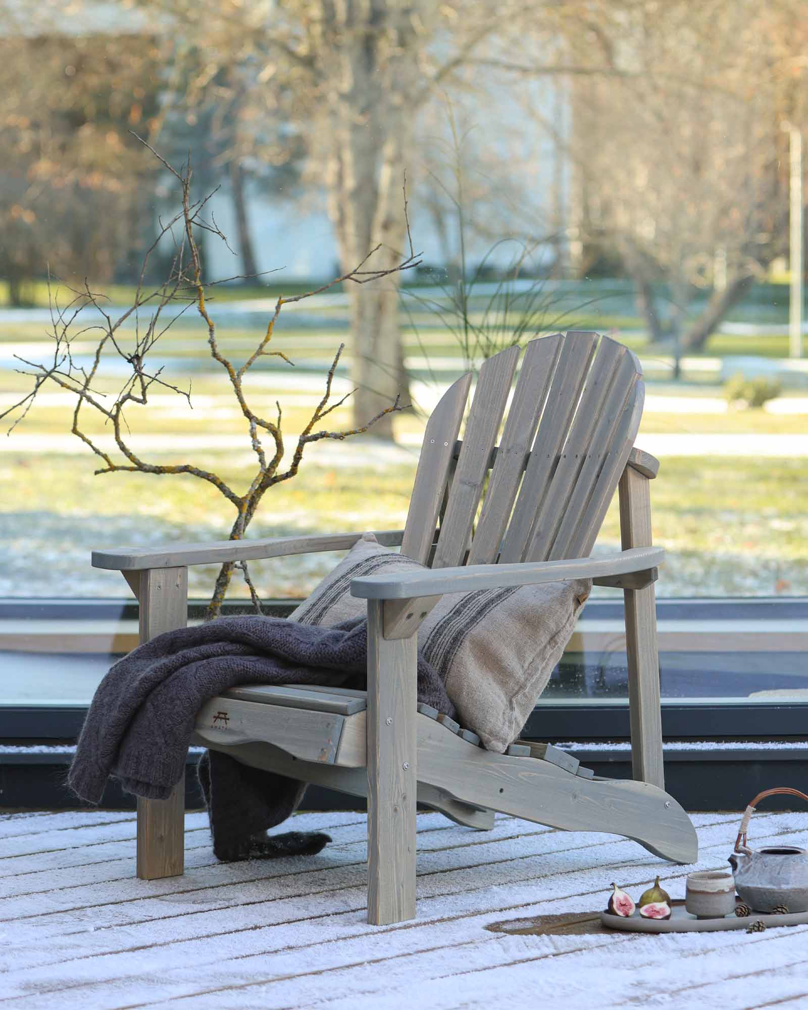 Grey wooden Adirondack chair with a decorative throw pillow and blanket on a snowy patio