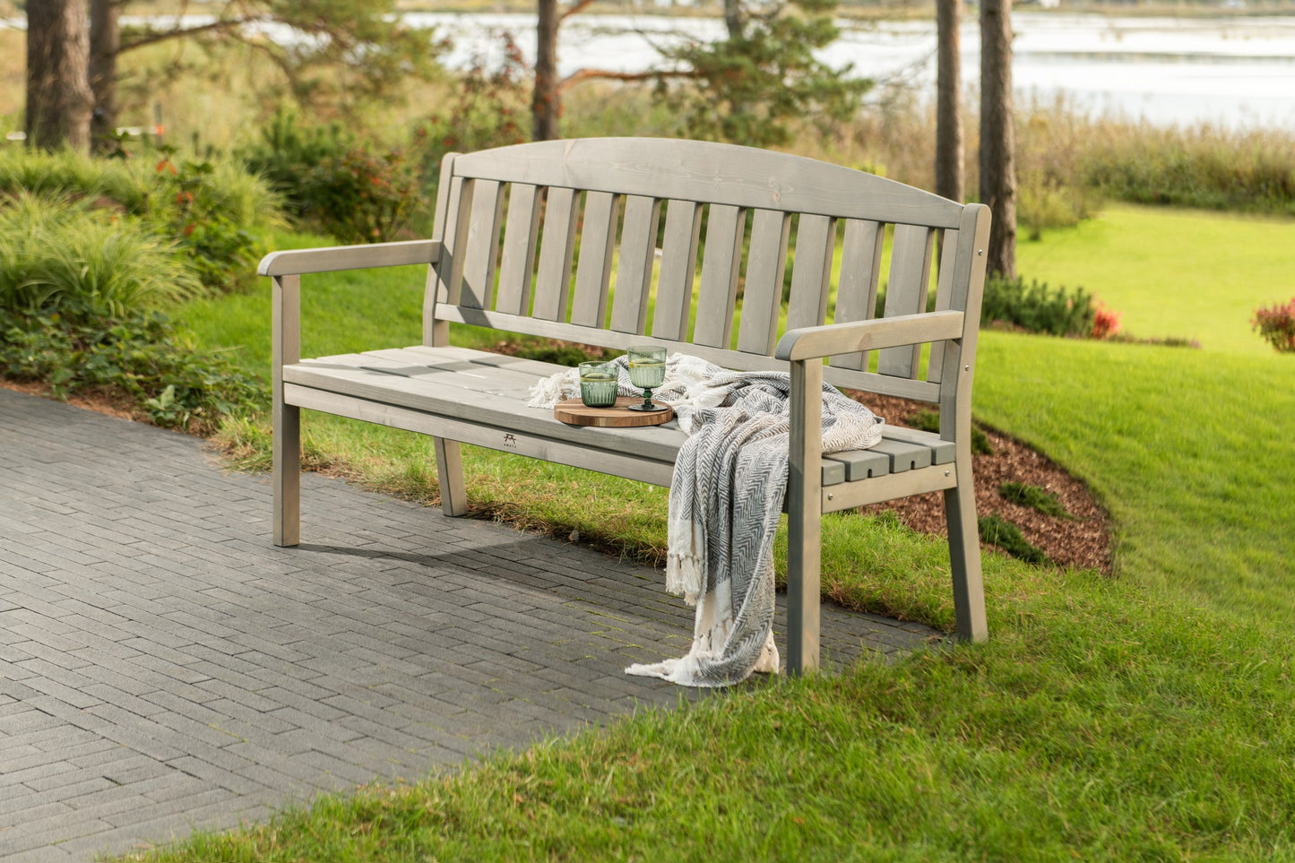 A light gray wooden garden bench with a curved backrest, situated on a paved path in a grassy park area with trees and water in the background.