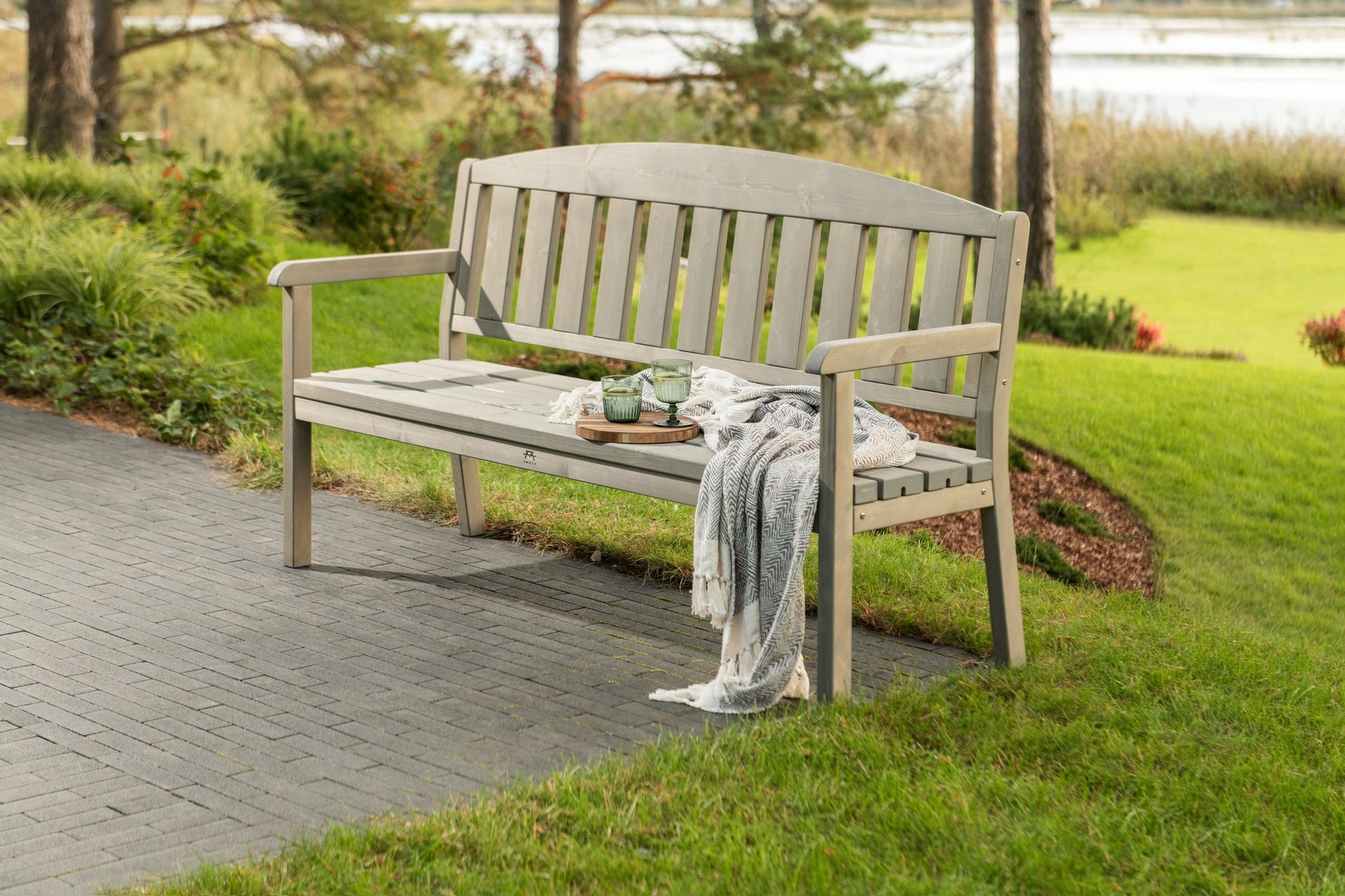 A light gray wooden garden bench with a curved backrest, situated on a paved path in a grassy park area with trees and water in the background.